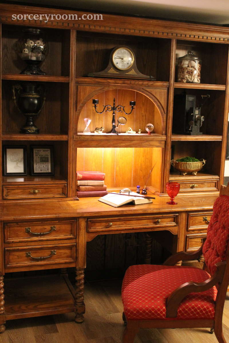 Red upholstered antique chair and a vintage desk with a hutch in a Harry Potter themed room with books, a magic wand, magnifying glass, red goblet, quill and ink, black candelier, glass orb, antique film projector, mantle clock, and a brass container with greenery.