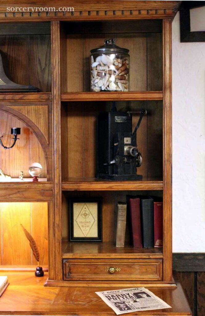 Accessories on a shelf in a Harry Potter themed room: glass jar with shells, antique film projector, quill and ink, old books, framed History of Magic book print