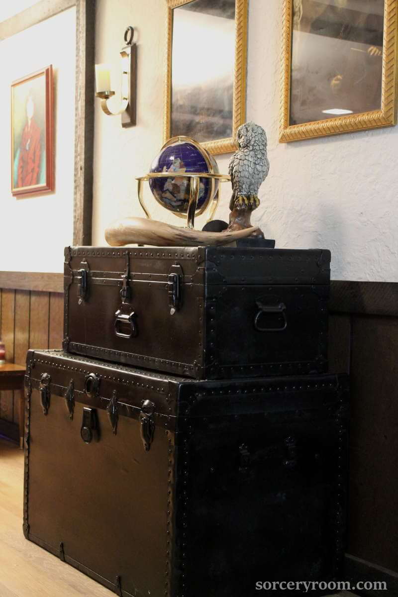 2 trunks painted black for a Harry Potter themed room, owl figurine, world globe, and a large horn on top, paintings in gold frames on the wall.