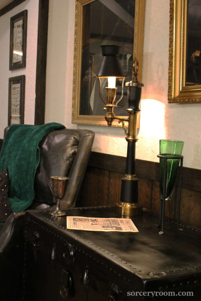 Slytherin themed sitting area with a black leather chair, green throw, black trunk, art on the walls, green vase, black and brass lamp, silver goblet, and a Harry Potter Undersirable No 1 paper print. 