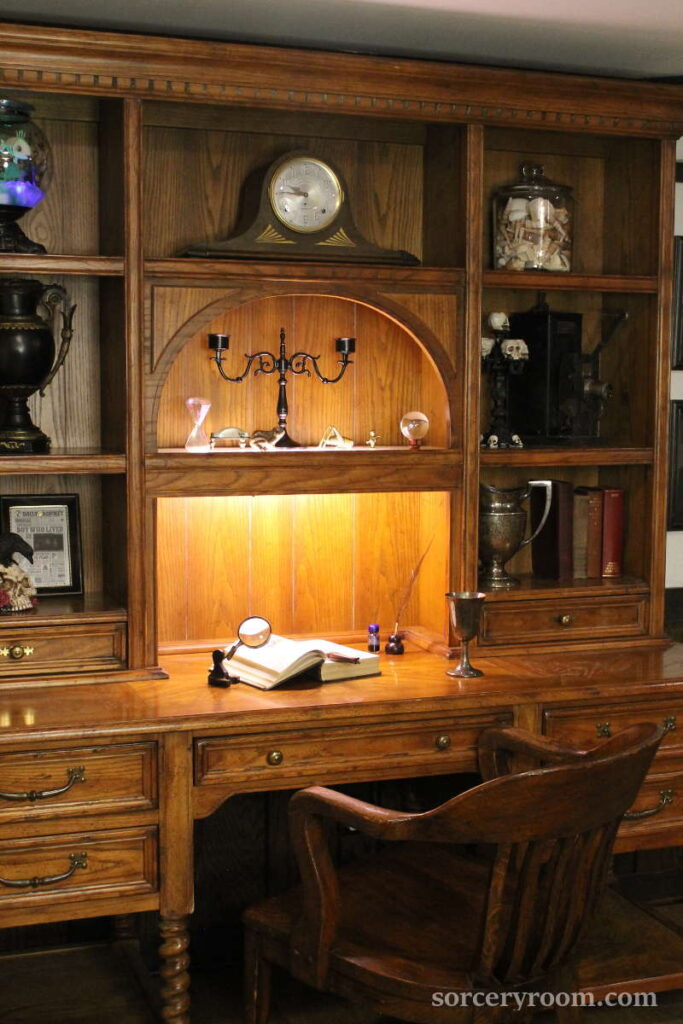 Vintage oak desk with a hatch used for a Harry Potter themed room. Accessories are a mantel clock, antique film projector, a glass jar with sea shells, old books. a silver goblet and a pitcher, magnifying glass, quill and ink 