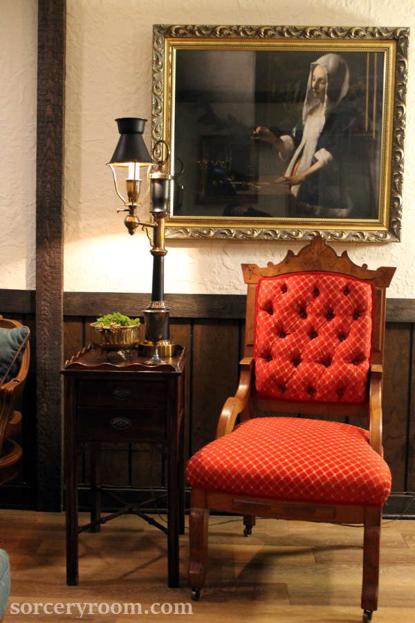 red upholstered chair, side table, brass vase with moss, silver goblet, lamp, gold frame