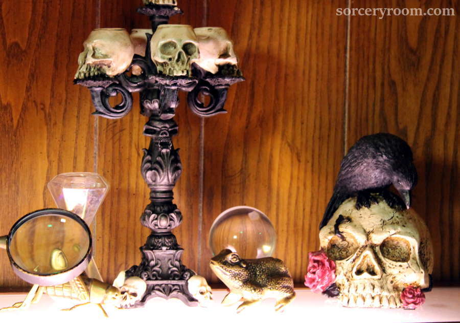 shelf with accessories: skull candelabra, magnifying glass, brass cricket, hourglass, brass frog, glass orb, skull with flowers and a raven on top