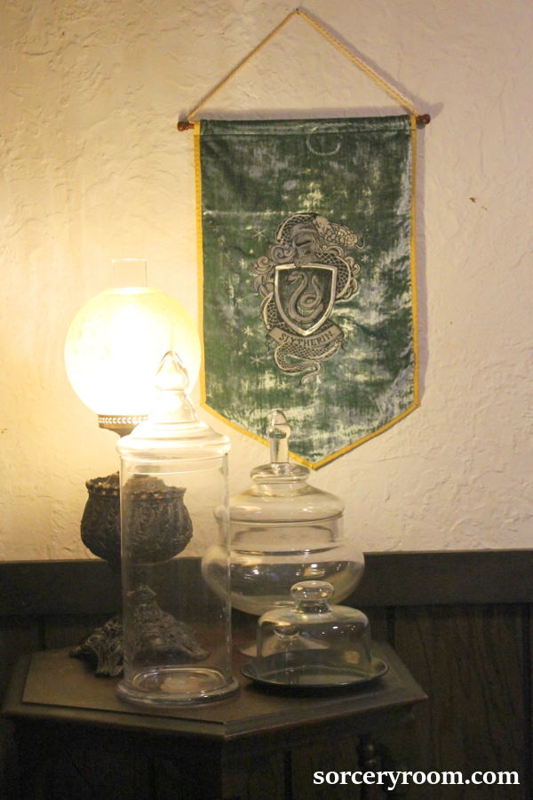 Slytherin pennant on the wall above a side table with a lamp and glass jars