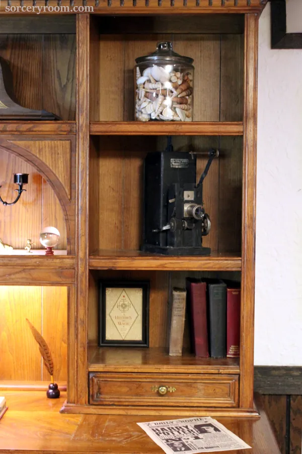 A wooden desk hutch holds a jar of seashells, an antique projector, and a few books. A Daily Prophet Harry Potter issue is on the desk.