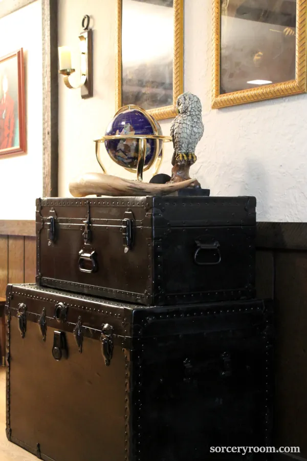 2 trunks painted black for a Harry Potter themed room, owl figurine, world globe, and a large horn on top, paintings in gold frames on the wall.