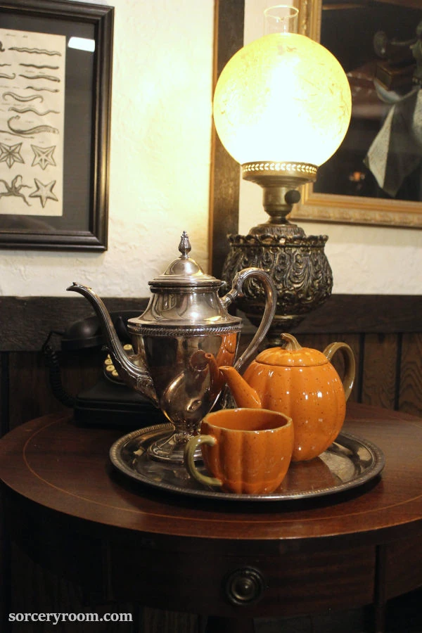 Harry Potter themed Halloween décor - a silver plated tea pot with a pumpkin shaped tea set on a table with an old lamp, black leather chair and old lamp in the background