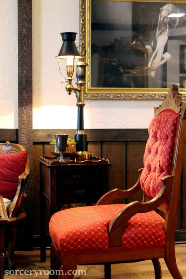 Harry Potter themed room with red upholstered chair, side table, brass vase with moss, silver goblet, brass and black lamp, painting in gold frame